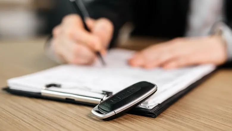 A black car key sits on a wooden desk, with a blurred person signing a document in the background.