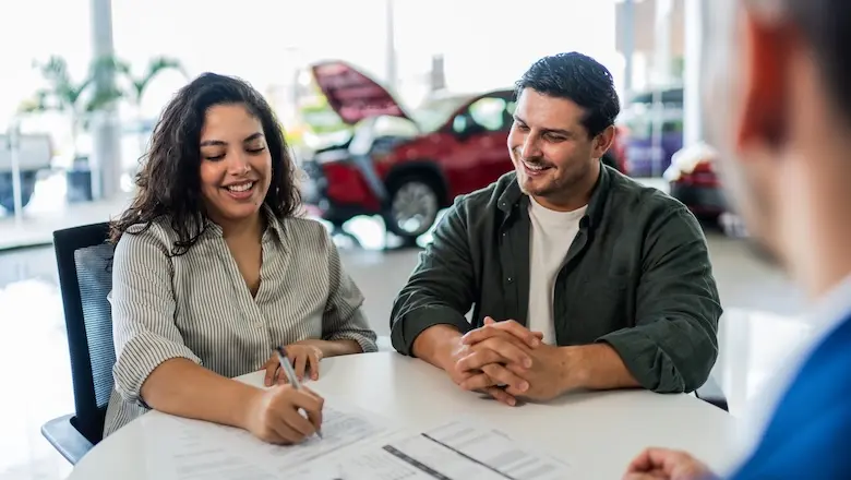 A man and a woman sit at a table in a car dealership with a car salesman while the woman signs a document.