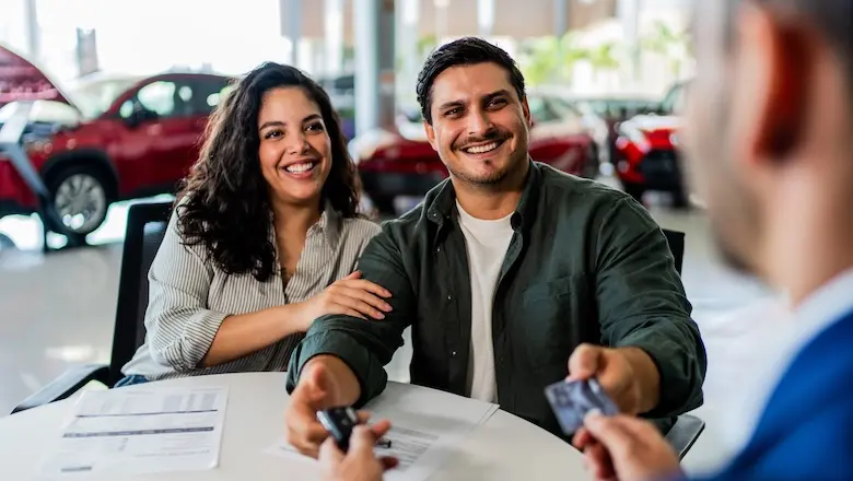 A man and a woman at a car dealership smile as the man hands his credit card to a salesman. The woman has her arm around the man.