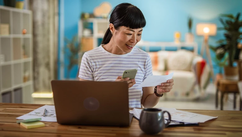 A woman reviewing a receipt on her phone and laptop while comparing budgeting options at a home desk.