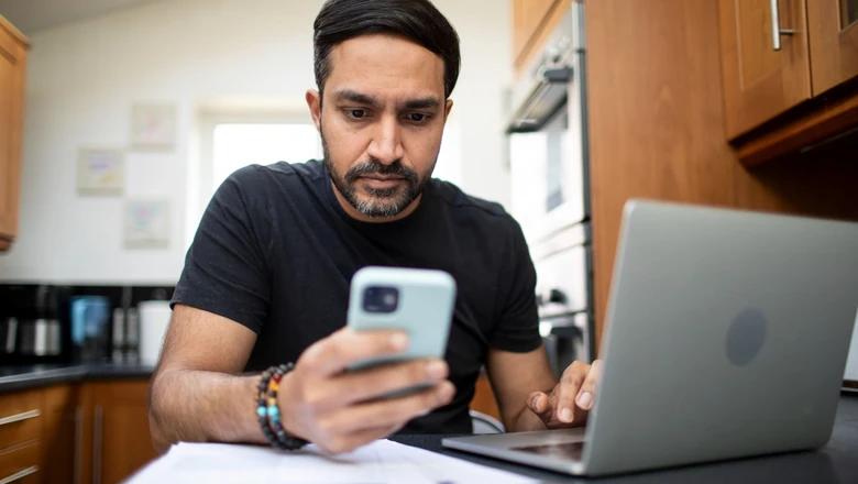 A man comparing budgeting apps on his phone and laptop at a kitchen counter with papers nearby.