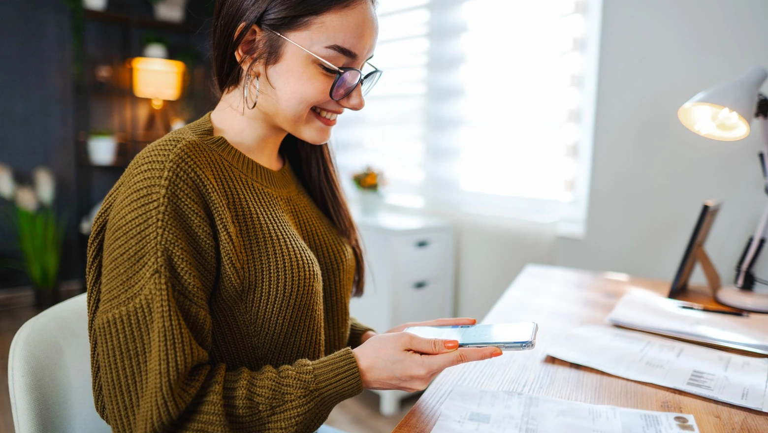 A woman in glasses photographs household bills with her phone while reviewing her budget at a home desk.