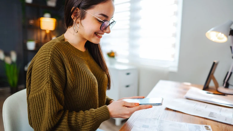 A woman in glasses photographs household bills with her phone while reviewing her budget at a home desk.