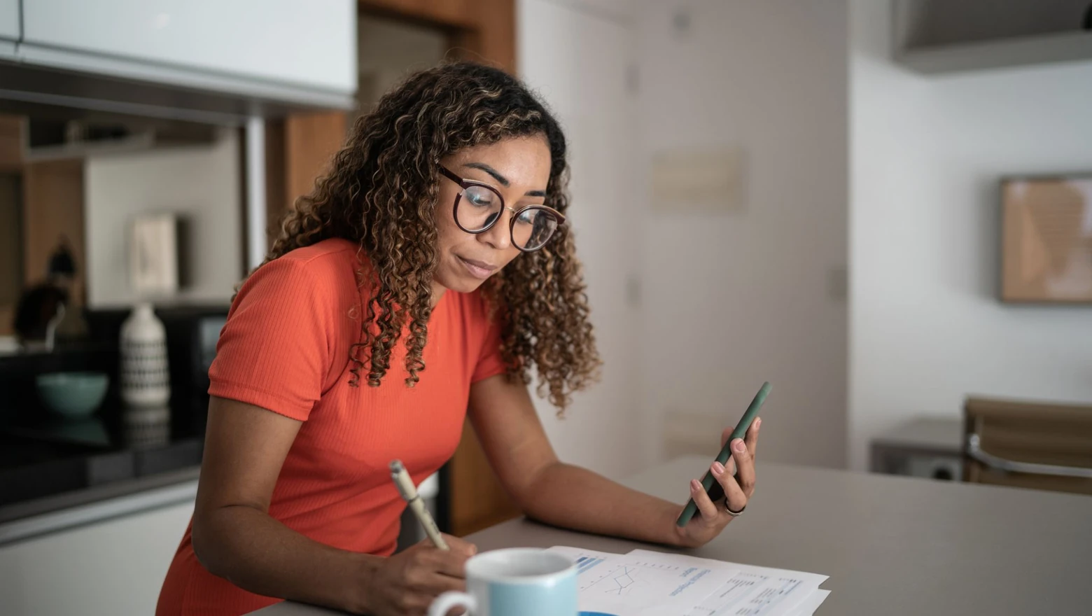 A woman in glasses reviews charts and her phone while comparing budgeting apps at a kitchen table.