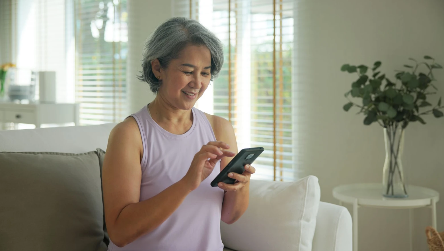 A woman smiling as she uses Coach Chat on her smartphone while seated on a sofa at home.