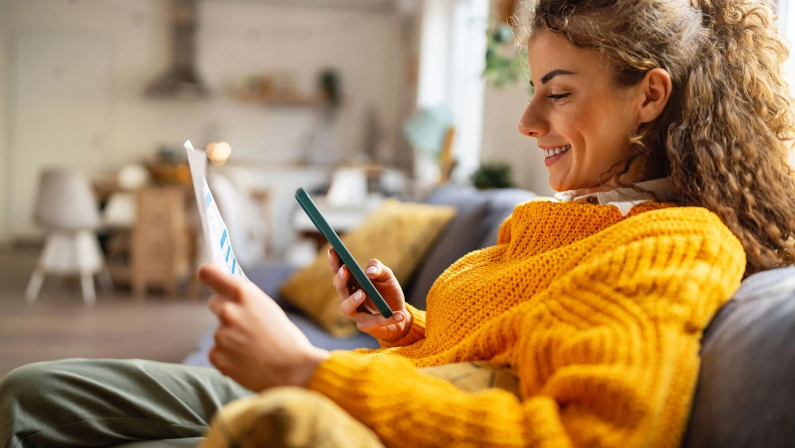 A woman smiles while comparing a financial report on paper to data on her smartphone.