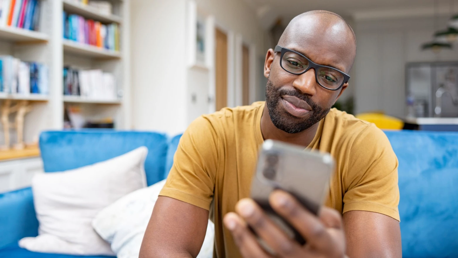 A man in glasses sits on a blue sofa reviewing a financial coaching app on his smartphone.
