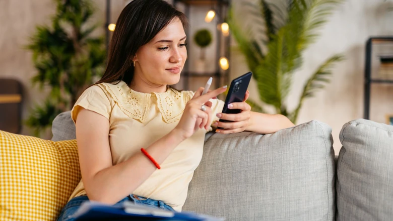 A woman on a sofa using her smartphone to explore signing up for SoFi Coach in a bright living room.