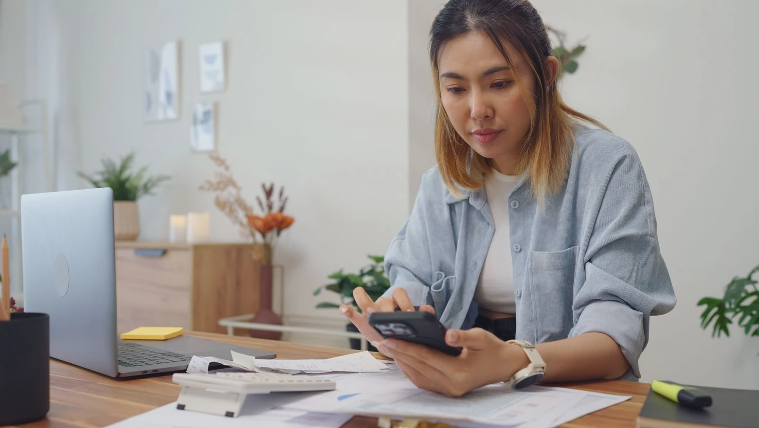 A woman uses a budgeting app on her phone while reviewing bills and paperwork at her desk.