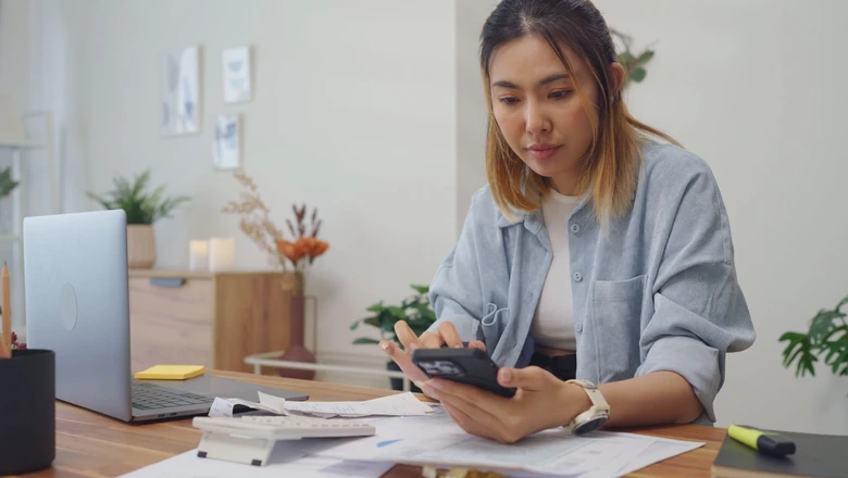 A woman uses a budgeting app on her phone while reviewing bills and paperwork at her desk.