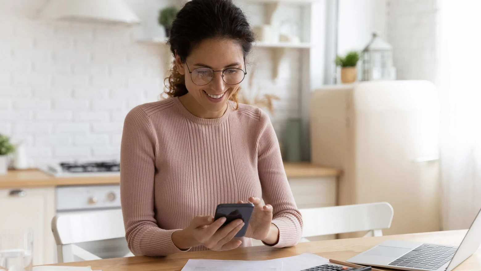 A woman in glasses reviews bills, a calculator, and her phone while comparing budgeting tools at home.