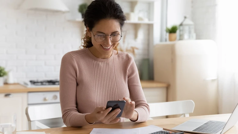 A woman in glasses reviews bills, a calculator, and her phone while comparing budgeting tools at home.