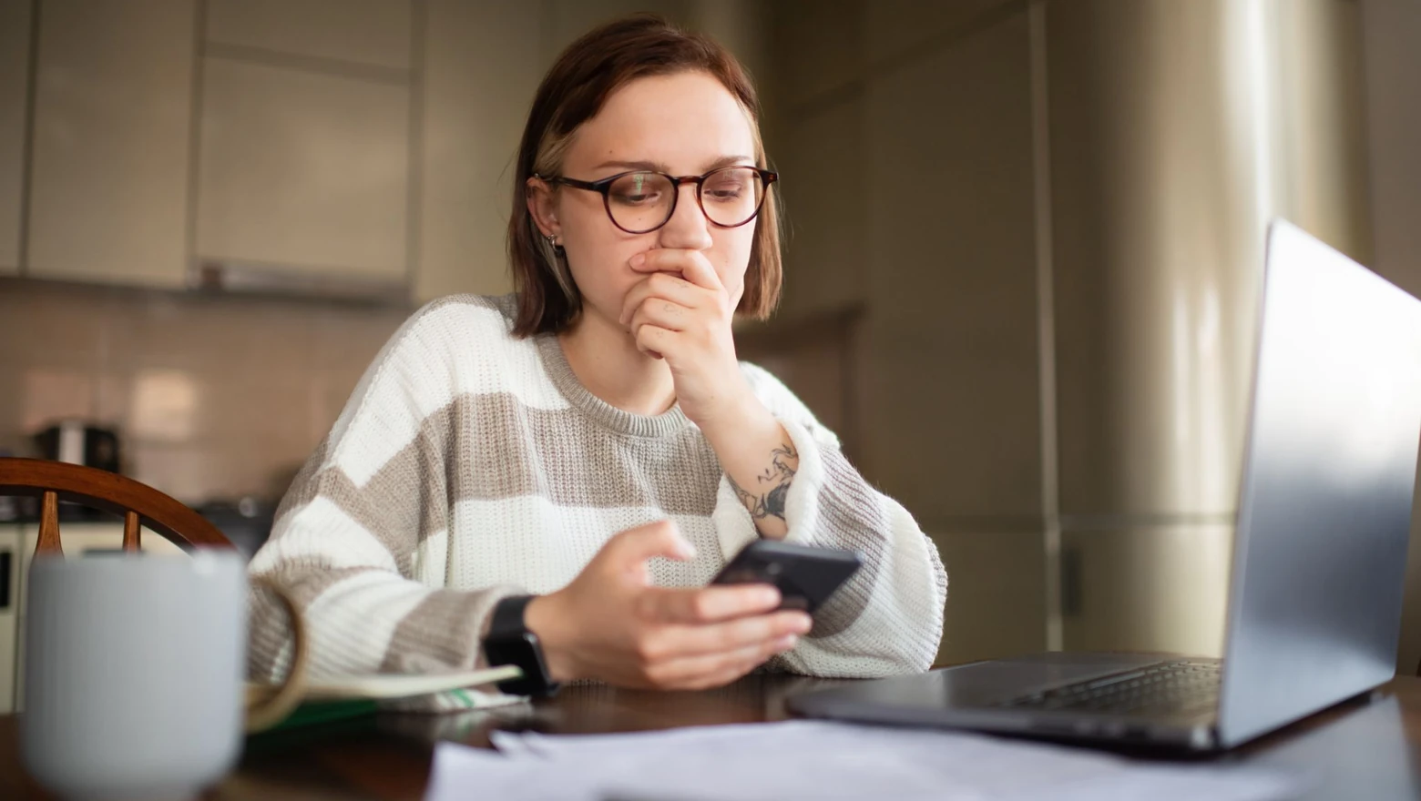 A woman in glasses studies her phone beside a laptop and papers while comparing budgeting apps at home.
