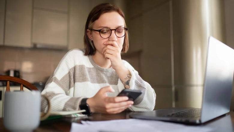 A woman in glasses studies her phone beside a laptop and papers while comparing budgeting apps at home.