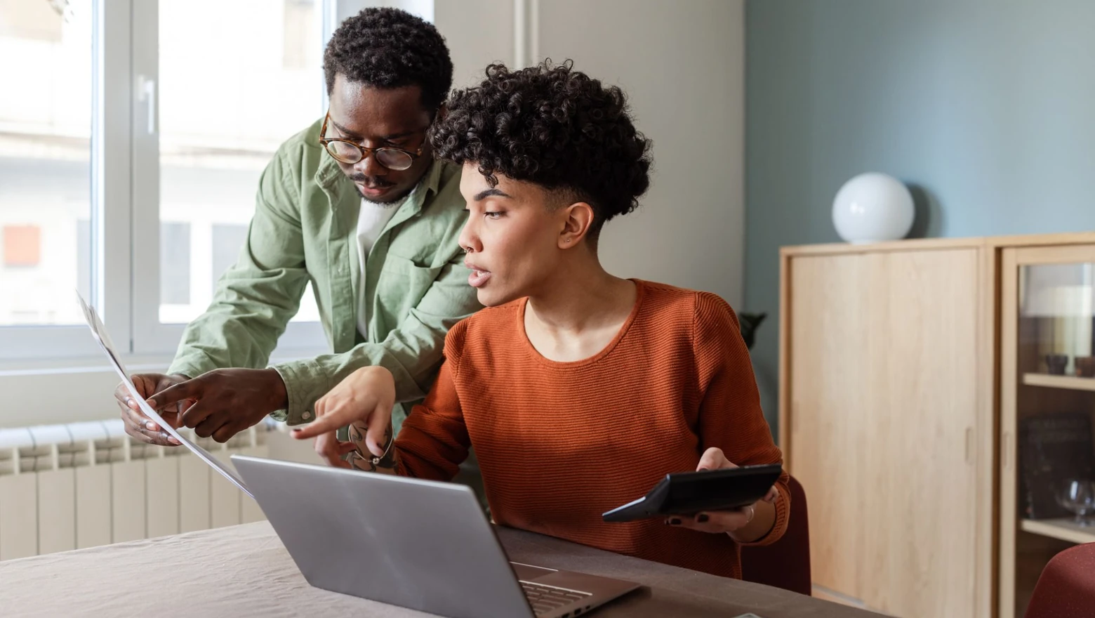 A couple reviews a budgeting comparison on a laptop at a table with papers and a calculator.