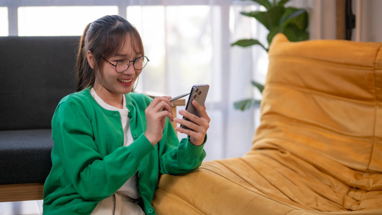 A smiling woman in glasses checks her credit score on her phone while seated in a bright living room.