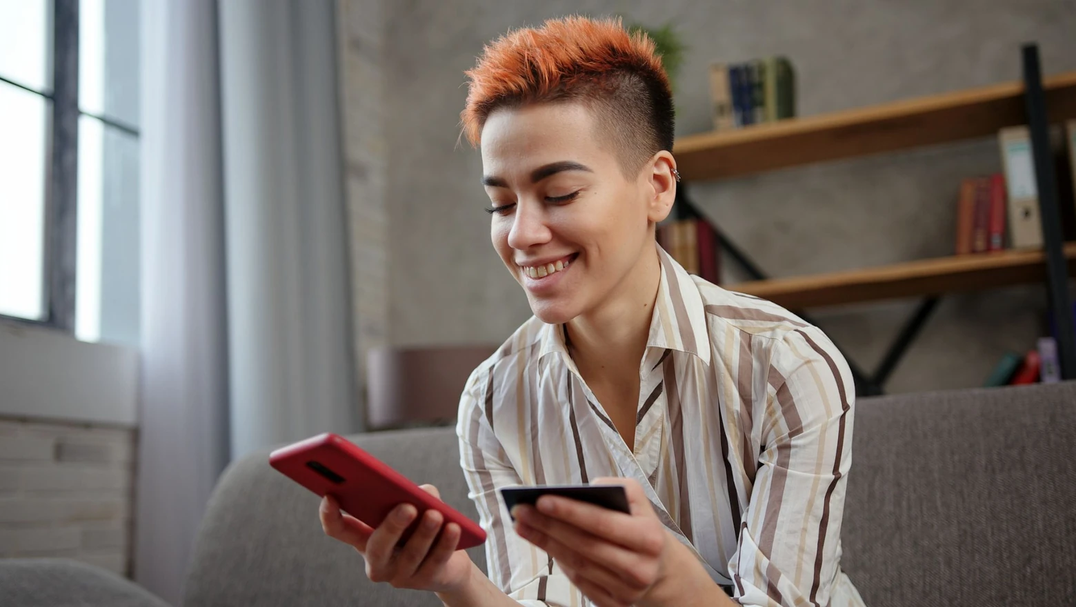 A smiling woman compares credit options using a smartphone and credit card while seated on a sofa.