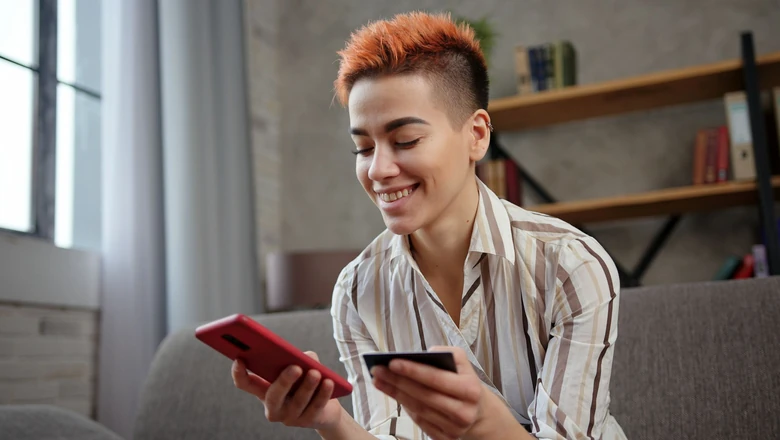 A smiling woman compares credit options using a smartphone and credit card while seated on a sofa.