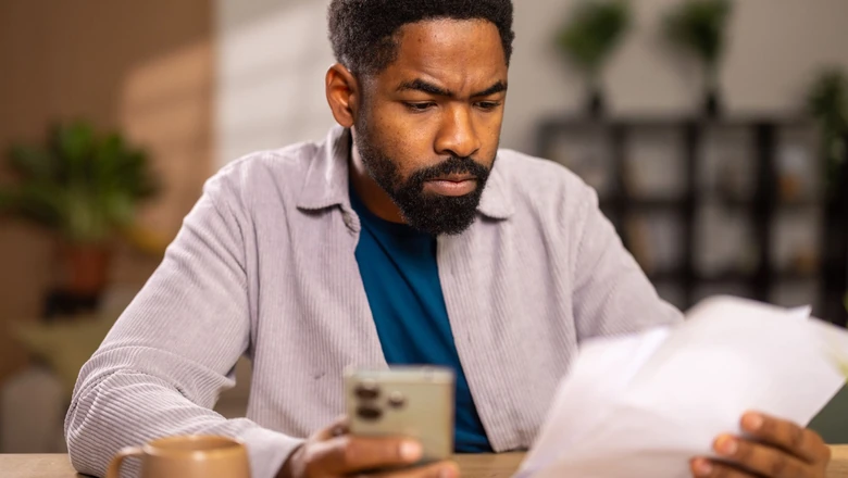 A man reviewing credit monitoring options on his phone and paper, comparing Aura and SoFi Coach.