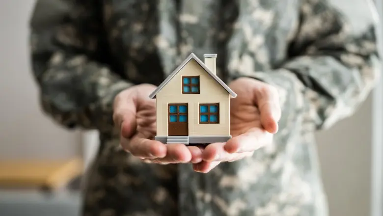 A person wearing military fatigues holds a small plastic model of a cream-colored house with brown and blue trim in both hands.