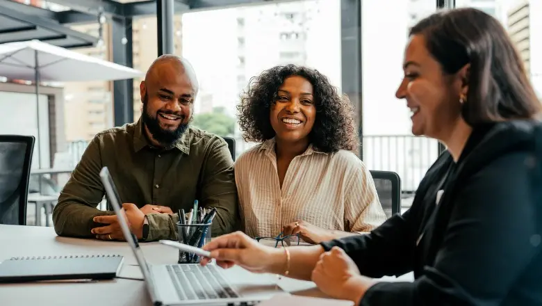 A smiling couple sits close together across the table from a woman who is looking at her computer screen and also smiling.