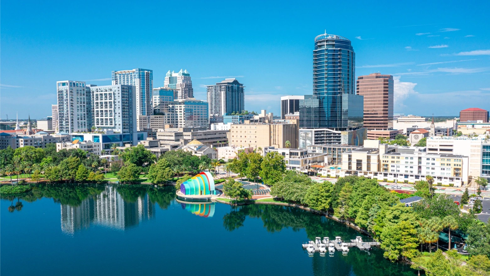 The skyline of downtown Orlando, Florida is reflected in the surface of a lake with the colorful sea-blue Walt Disney Amphitheater at center.