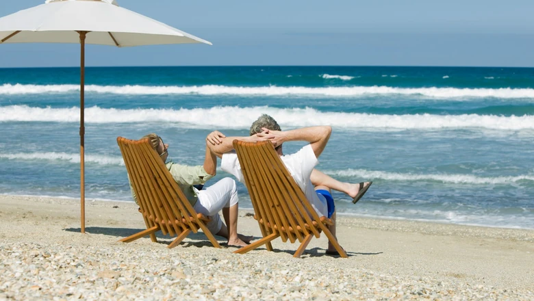 A retired couple sitting on chairs at the beach near an umbrella.