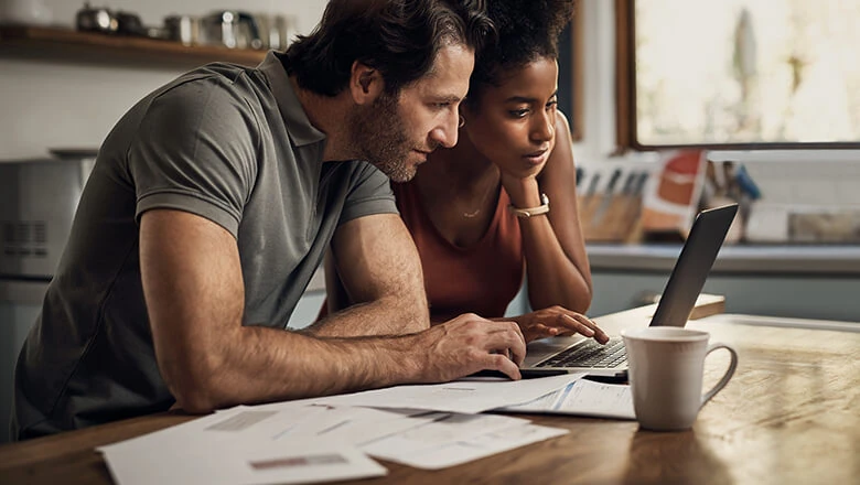 A couple reviews financial documents and a laptop at a kitchen table, researching 401(k) tax rules.