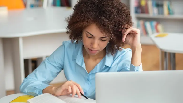 A graduate student reviewing a textbook while working on a laptop at a desk in a school setting.