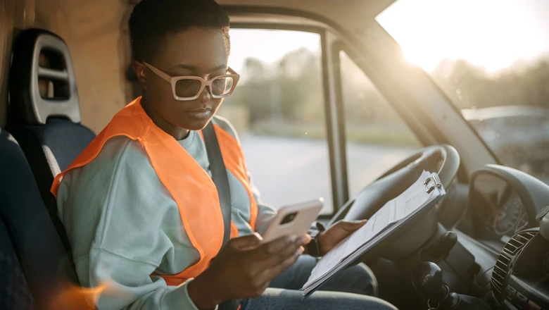 A gig worker in an orange safety vest sits in a delivery van reviewing a clipboard and using her phone.