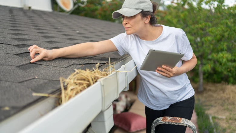 A woman on a ladder inspecting the roof of her second home while holding a tablet.
