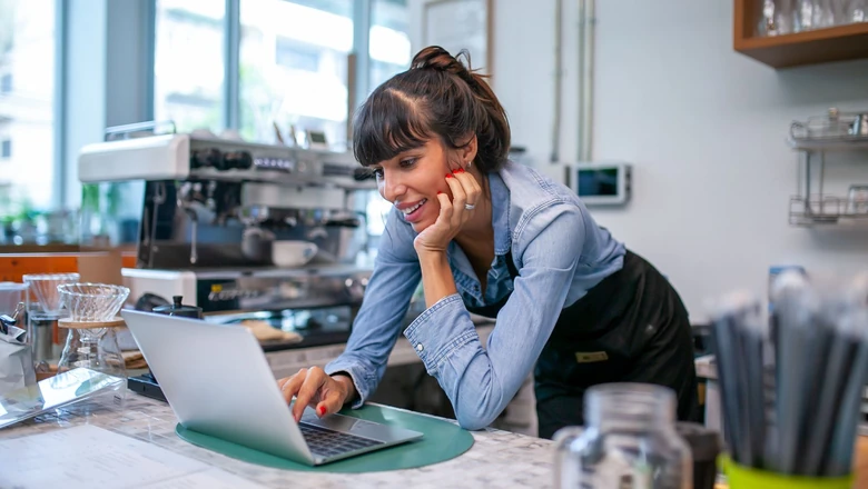 A small business owner at a counter using a laptop to research the difference between an invoice and a bill.