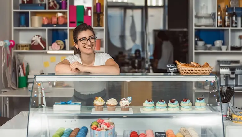 A small business proprietor smiling behind a counter showcasing cupcakes and other pastries