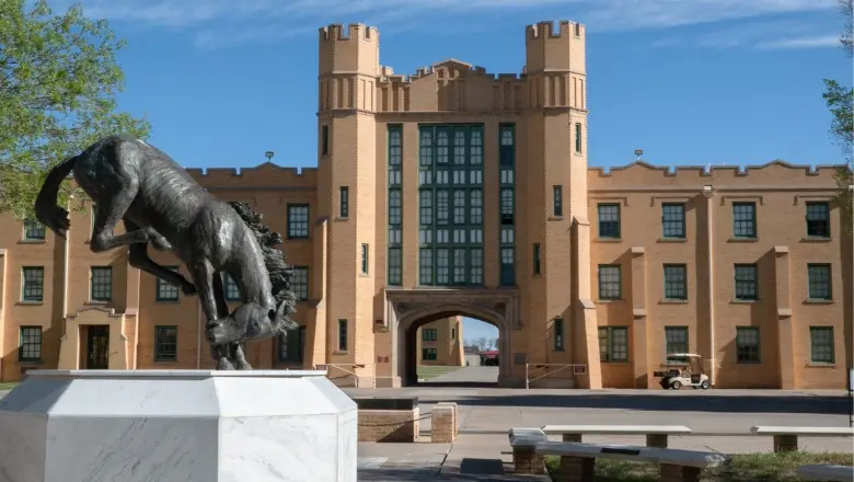 A statue of a bucking horse stands in front of a tan brick military building with a large archway.