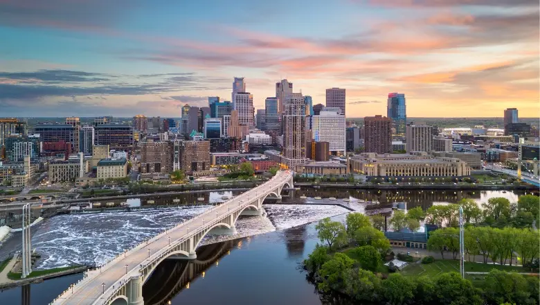 An aerial view of the Minneapolis skyline at sunset, with a bridge over the Mississippi River in the foreground.