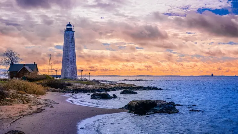 A scenic shot of the New Haven Lighthouse on a rocky shore, with a colorful sunset in the distance.