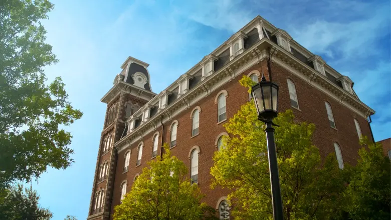 A low-angle view of a large, historic brick building with a pointed roof, surrounded by green trees.