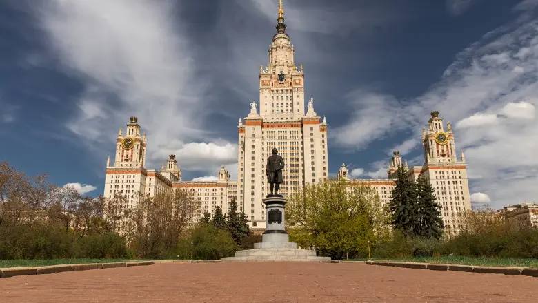 A grand, historic university building with a central spire and side towers, with a statue in the foreground.